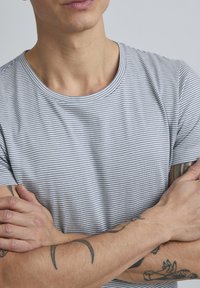 Harmaa ja valkoinen raidallinen t-paita, jossa on pyöreä kaula-aukko ja lyhyet hihat, valmistettu pehmeästä kankaasta. Käsiin näkyviä tatuointeja, kädet ristissä.