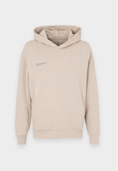 Bež hoodie z sprednjim kenguru žepom, dolgimi rokavi in kapuco ter diskretno besedilno podrobnostjo na prsih. Mehka, gladka tekstura tkanine.