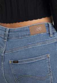 Primer plano de un bolsillo trasero y la cinturilla de unos vaqueros Lee azules con parche de piel de la marca Lee, combinados con un top de punto negro recortado.