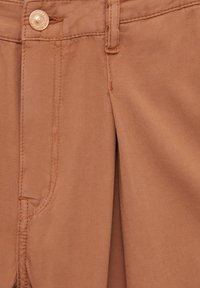 Tissu de pantalon marron en gros plan montrant un bouton, une passants de ceinture, une poche avant et des détails de couture.