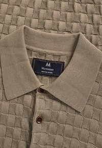Polo beige en maille avec motif damier texturé, boutons marron et étiquette "Matinique Heritage Apparel" à l'intérieur du col.