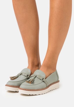 Mocassins en cuir vert clair avec des franges et de épais semelles blanches portées pieds nus sur fond blanc.