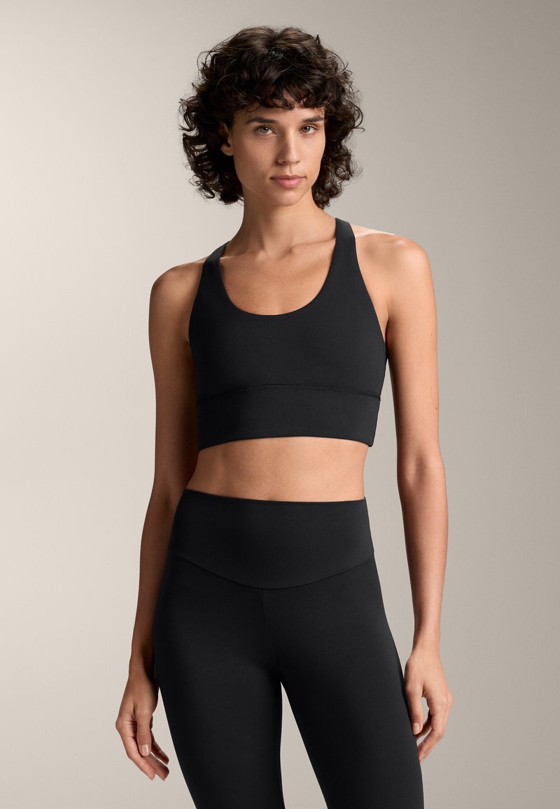 Reggiseno sportivo nero con scollatura a barchetta e spalline larghe, abbinato a leggings neri a vita alta. Tessuto dalla consistenza liscia, design minimalista.