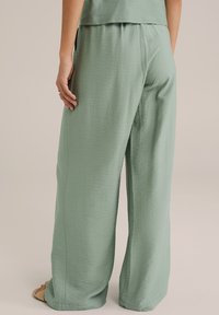 Ljust gröna, vida byxor i textil med struktur. Elastisk midja och sidofickor synliga. Komplementeras av neutrala sandaler.