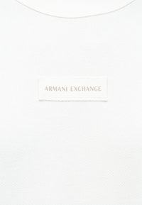 Λευκό ύφασμα με μια μικρή ορθογώνια ετικέτα Armani Exchange ραμμένη στο κέντρο, που εμφανίζει το όνομα της μάρκας σε ανοιχτό γκρίζο χρώμα.