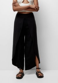 Pantalon palazzo en lin noir avec fentes latérales, doté d'une taille haute et d'une coupe décontractée. Associé à des sandales noires à fines lanières.