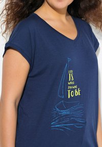 Marineblå T-shirt med korte ærmer og V-hals, med et grafik af en sejlbåd og teksten "Livet er, hvor du ønsker at være" i gul.