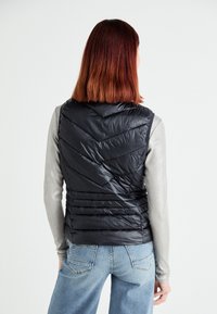 Black puffer vest with chevron quilted pattern, high collar, and zippered front. Paired with a long-sleeve silver top and denim jeans.