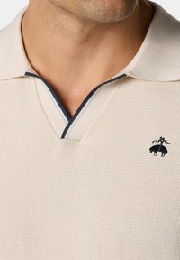 Beige Polo-Shirt mit strukturiertem Strick, versehen mit einem marineblauen Akzent am Kragen und einem kleinen schwarzen gestickten Logo auf der Brust.