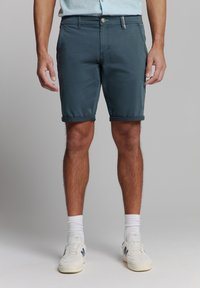 Heren shorts met opgerolde manchetten in donker teal, gemaakt van lichte stof. Voorzien van een voorste knoopsluiting en zijzakken. Gecombineerd met witte sneakers.