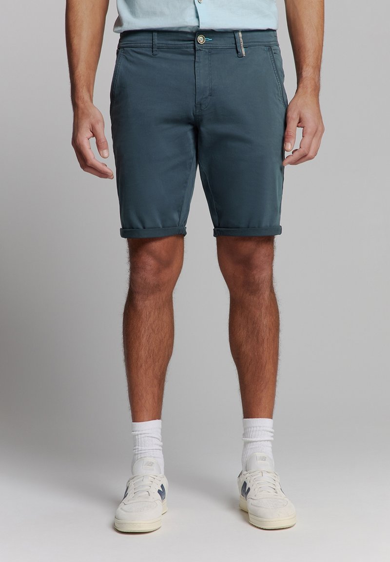 Heren shorts met opgerolde manchetten in donker teal, gemaakt van lichte stof. Voorzien van een voorste knoopsluiting en zijzakken. Gecombineerd met witte sneakers.