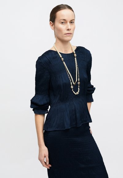 Femeie purtând o bluză texturată de culoare bleumarin închis, cu mâneci bufante și o fustă asortată, accesorizată cu un colier lung de perle.