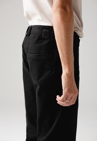 Μαύρο βαμβακερό παντελόνι με στενή γραμμή, που διαθέτει πίσω τσέπη και ανάγλυφη ετικέτα στη μέση. Το χέρι τοποθετημένο κοντά στο ισχίο.