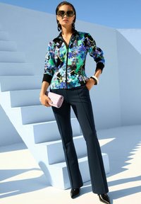 Vrouw in een bloemen-ritsjack, marineblauwe flared broek, zwarte puntige schoenen, groene zonnebril, met een kleine roze clutch tegen blauwe trappen en de lucht.