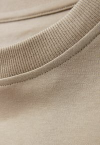 Beige bavlněná tričkovina v detailu, se hladkou texturou a žebrovaným výstřihem, ukazující jemné švy a hladký povrch.
