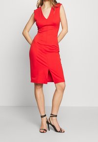 Červené upnuté šaty s výstřihem do V, krátkými rukávy a předním rozparkem. Hladká textura, délka po kolena, doplněné černými páskovými podpatky.