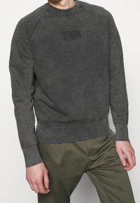 Szara bluza wykonana z miękkiego materiału, z okrągłym dekoltem, rękawami raglanowymi oraz wytłoczonym logo na środku klatki piersiowej. Gładka faktura.