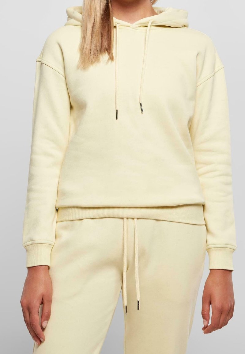 Femme portant un sweat à capuche jaune clair et un pantalon de survêtement assorti avec des cordons, montrée des épaules aux hanches sur un fond uni.
