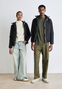 Schwarze Jacken mit Reißverschluss und elastischen Bündchen, kombiniert mit einem cremefarbenen Rippsweater, hellblauen weit geschnittenen Jeans und olivgrünen Hosen. Weiße Sneaker.