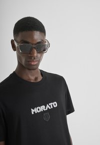 Camiseta de algodón negra con el texto blanco "MORATO" y un emblema de oso negro. Modelo que lleva gafas de sol geométricas grises transparentes.