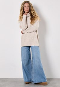 Beżowy sweter dzianinowy z kołnierzem, połączony z jasnoniebieskimi szerokimi spodniami dżinsowymi i brązowymi butami na platformie na szarym podłożu.