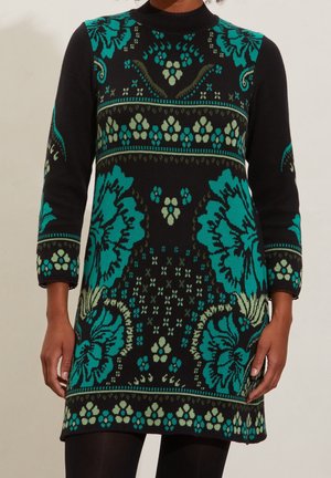 Zwarte lange mouwen jurk met grote bloemenpatronen in teal en lichtgroen en banden met kleine bloemen, gedragen met zwarte panty's.