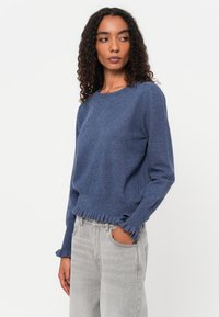 Sweater azul com padrão de diamante, decote redondo e punhos com babados. Combinado com jeans cinza claro, com um corte relaxado.