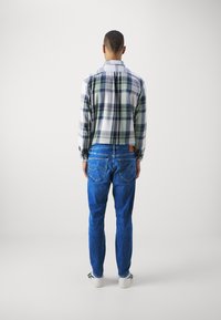 Camisa de cuadros para hombre en tonos verdes y azules, combinada con jeans de mezclilla azules. Zapatillas blancas con detalles en negro visibles en los pies.