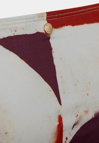 Tkanina s patterni, ki prikazuje geometrijske oblike v burgundski, rjavi in kremni barvi. Na šivu je vidna ikona iz zlatega kovinskega materiala. Mehka tekstura.