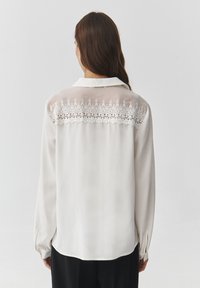 Blusa blanca de manga larga hecha de una tela ligera, con un respaldo transparente y un delicado bordado floral en los hombros.