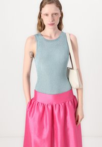 Top in morbido verde menta a costine abbinato a una gonna di raso rosa con una vita ampia. La modella tiene una piccola borsa bianca, lo sfondo è bianco uniforme.