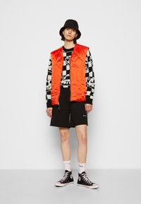 Colete acolchoado, de cor laranja brilhante, sobre um suéter com padrão preto e branco, combinado com calções pretos e ténis de cano alto pretos.