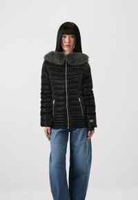 Svart pufferjakke med fuskepelskrage, med gullglidelåser og horisontal søm, kombinert med løstsittende blå jeans.