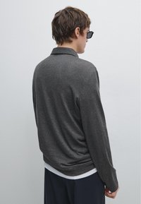 Dunkelgraues Strickhemd mit langen Ärmeln und umgeschlagenem Kragen, im lässigen Schnitt. Über einem weißen Hemd getragen, kombiniert mit dunklen Hosen und Sonnenbrille.