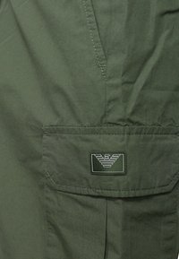 Giacca verde oliva in tessuto liscio, con taschino applicato e etichetta con logo raffigurante ali stilizzate. Design semplice e funzionale.