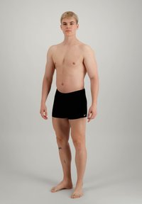 Sorte badebukser laget av elastisk stoff, med en glatt tekstur og en liten hvit Nike-logo foran. Modellen står barbent.