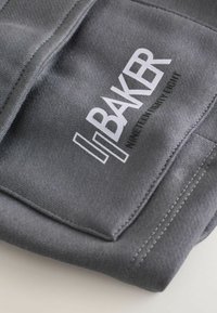 Gros plan sur un tissu gris avec une poche cousue affichant le logo blanc "BAKER" et un texte plus petit indiquant "NINETEEN EIGHTY LIGHT".