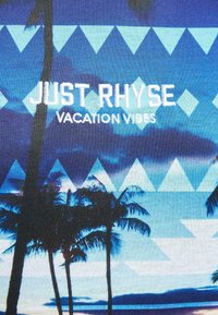 Marškinėliai su mėlynu gradientu, palmės siluetais ir trikampio raštais. Balti žodžiai skelbia "JUST RHYSE" ir "VACATION VIBES."