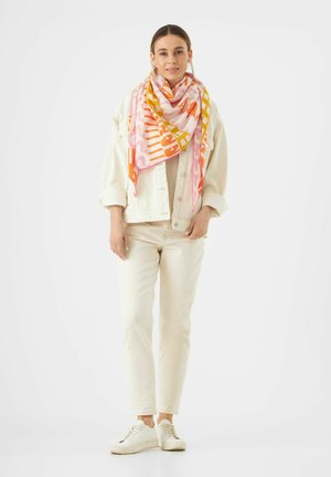Écharpe colorée à motifs dans des tons de rose, orange et jaune, enroulée autour du cou. Le modèle porte une veste crème et un pantalon avec des chaussures blanches.