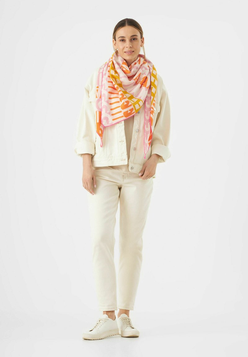 Écharpe colorée à motifs dans des tons de rose, orange et jaune, enroulée autour du cou. Le modèle porte une veste crème et un pantalon avec des chaussures blanches.