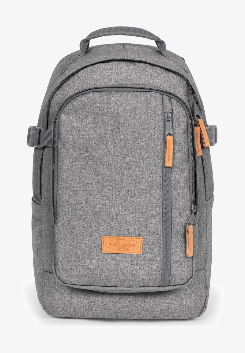 Sac à dos en tissu gris avec une finition texturée, doté d'une étiquette en cuir tan, de poches zippées et de bretelles réglables.