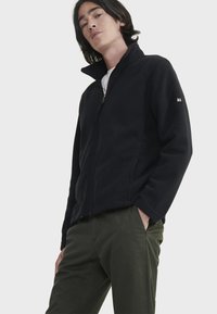 Schwarze Fleecejacke mit hohem Kragen, vorne mit Reißverschluss und seitlichen Taschen. Verfügt über dezentes Branding am Ärmel. Einfaches, klassisches Design.