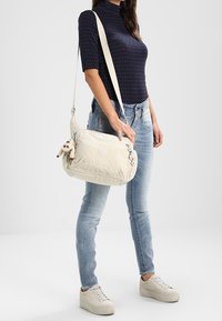 Borsa a spalla di un beige chiaro con una finitura testurizzata, dotata di un logo rotondo, tracolla regolabile e un piccolo accento in peluche attaccato.