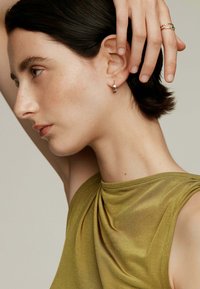 Pendientes de aro de oro con acentos circulares, usados por una persona con una blusa verde sin mangas y con escote drapeado. Textura de piel suave visible.