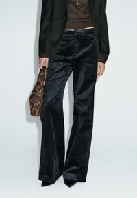 Pantaloni in corduroy a zampa di elefante di un blu scuro, con una superficie testurizzata. Abbinati a un blazer nero e con una borsa in pelle serpente.