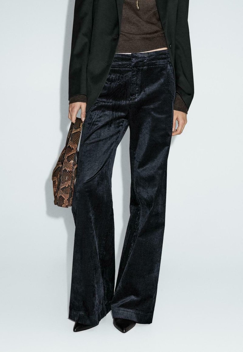 Pantaloni in corduroy a zampa di elefante di un blu scuro, con una superficie testurizzata. Abbinati a un blazer nero e con una borsa in pelle serpente.