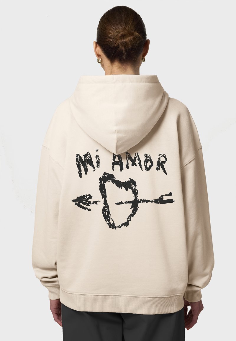 Beige oversize kapuce ar stringiem. Mugurpusē liela melna grafika ar sirdi un bultu, apzīmēta "Mi Amor". Ribotās aproces.