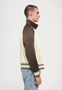 Lichte zip-up jas met crème en bruine panelen, geribde manchetten en zoom, met subtiele gestreepte accenten. Soepele textuur en moderne pasvorm.