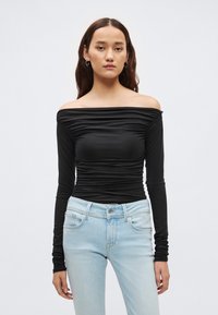 Zwarte off-shoulder top met lange mouwen en gerimpelde textuur; gecombineerd met lichtblauwe jeans met een klassieke vijfzakjesontwerp.