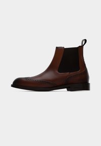 Bottes Chelsea en cuir marron avec panneaux latéraux élastiques, détails à brogue le long du bout et une languette à l'arrière. Semelle en caoutchouc plate.